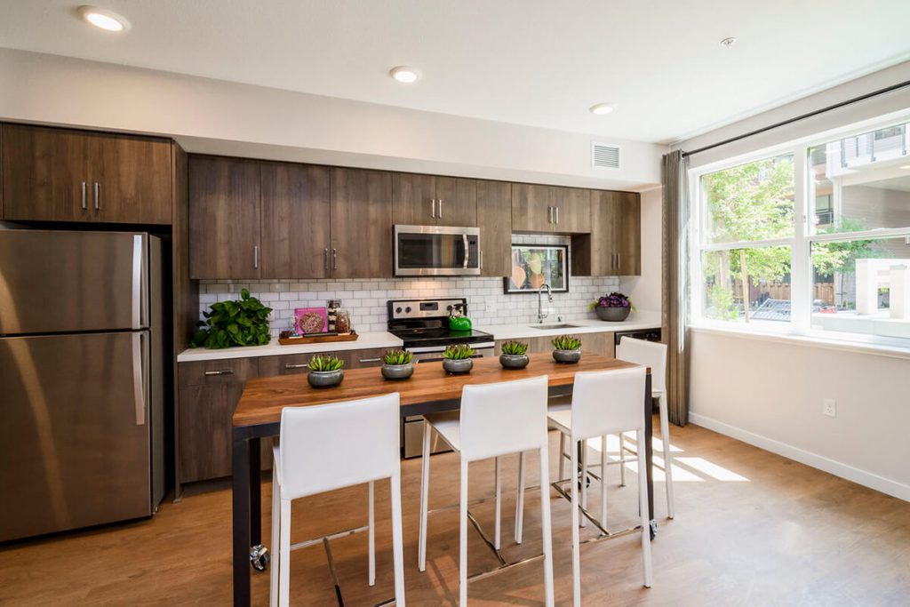 Apartments in Sunnyvale, CA - 6TenEAST - Kitchen with Hardwood-Style Plank Flooring, Stainless Steel Appliances, Brown Wood Cabinetry, White Quartz Countertops, Tile Backsplash, Large Window, and Decorative Plants