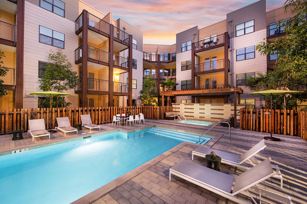 Luxury Apartments in Sunnyvale, CA - 6TenEAST - Gated Pool Area with Lounge Chairs, Umbrellas, Hot Tub, Trees, and View of Building Exterior